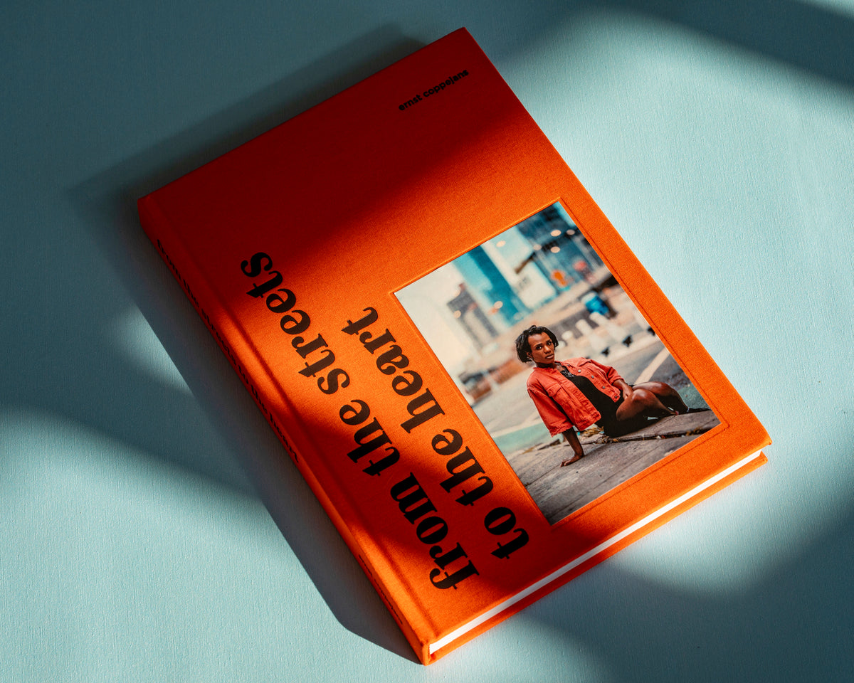 Hardcover photobook From the Streets to the Heart by Ernst Coppejans, orange cloth cover with a portrait, lying in the sun on a light blue surface